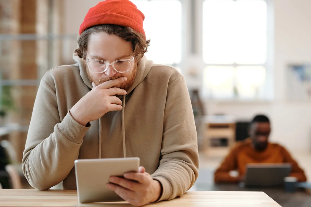 SEO operator in a red beanie reading on a tablet at a coworking desk, colleague on a laptop in the background — choosing the right tools for SEO in 2026